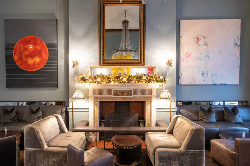 Festive fireplace with garland and Eiffel Tower mirror in Home House lounge, gray sofas, abstract art, warm lighting.