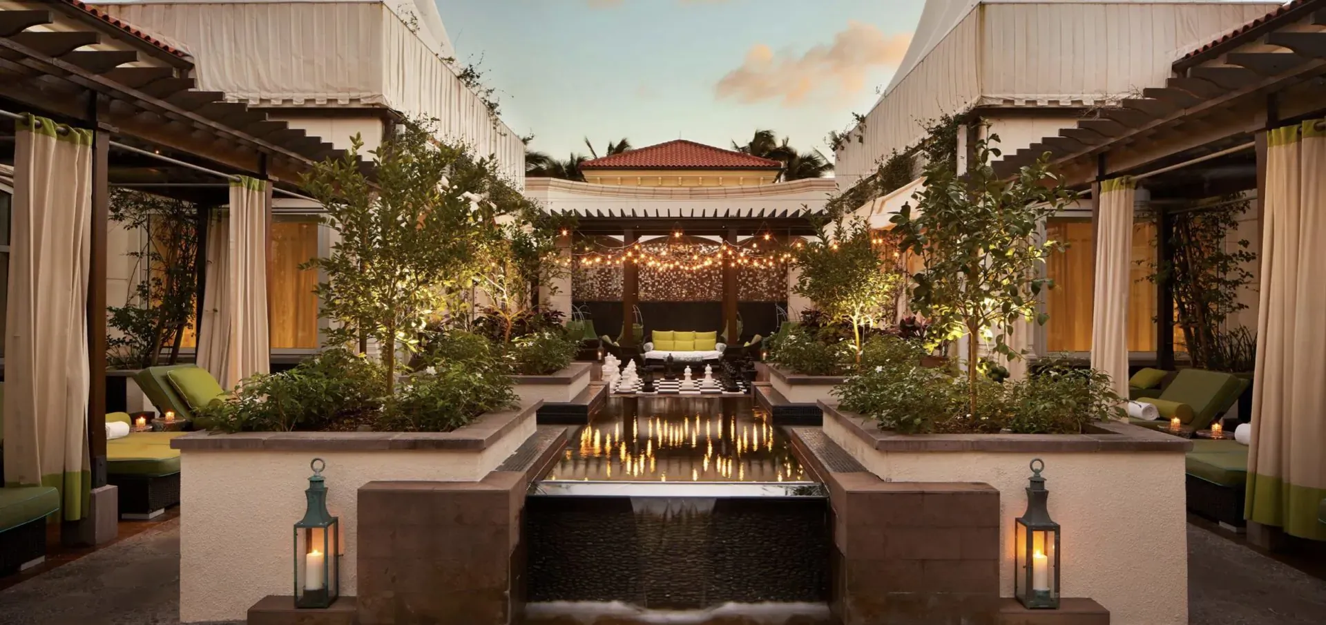 Luxurious outdoor courtyard at dusk with central fountain, lounge seating, lanterns, plants, and pergola drapes