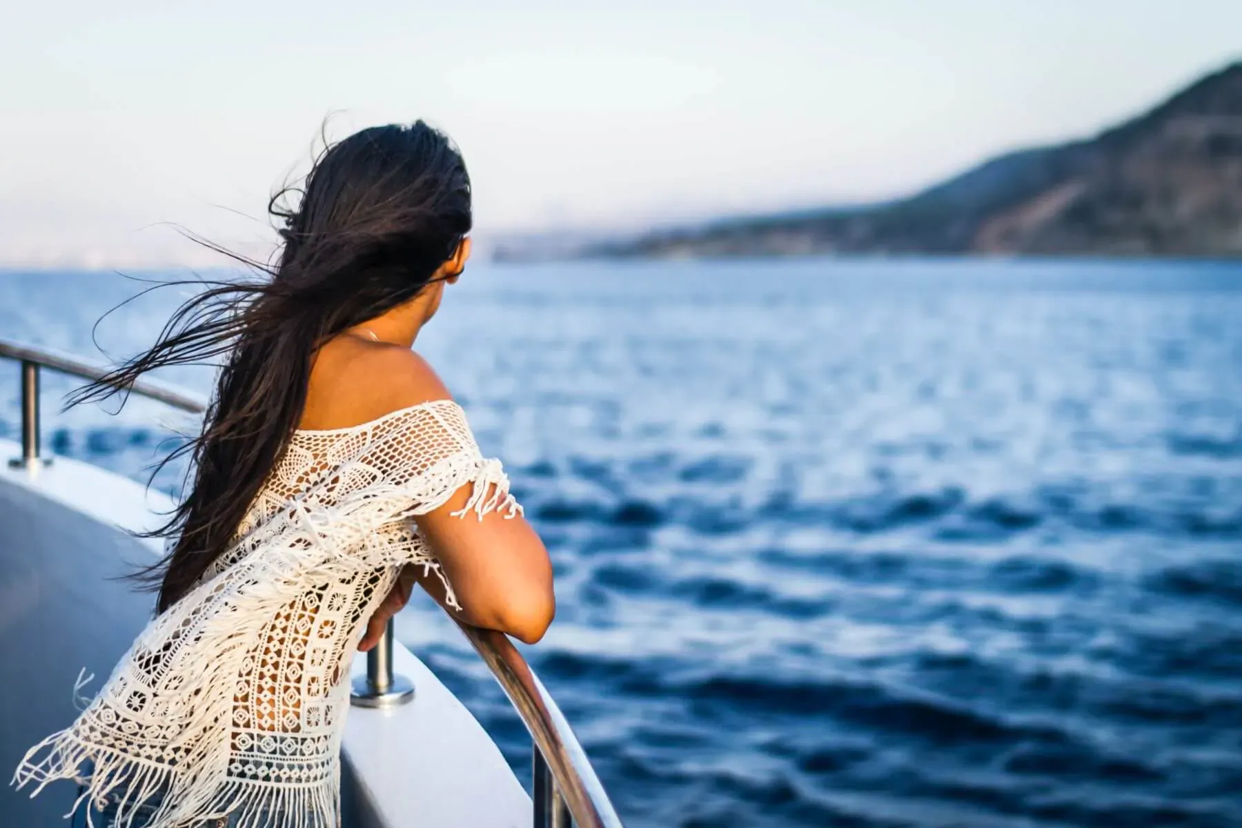 Woman in white lace dress stands on yacht railing, gazing at sea and mountains at dusk