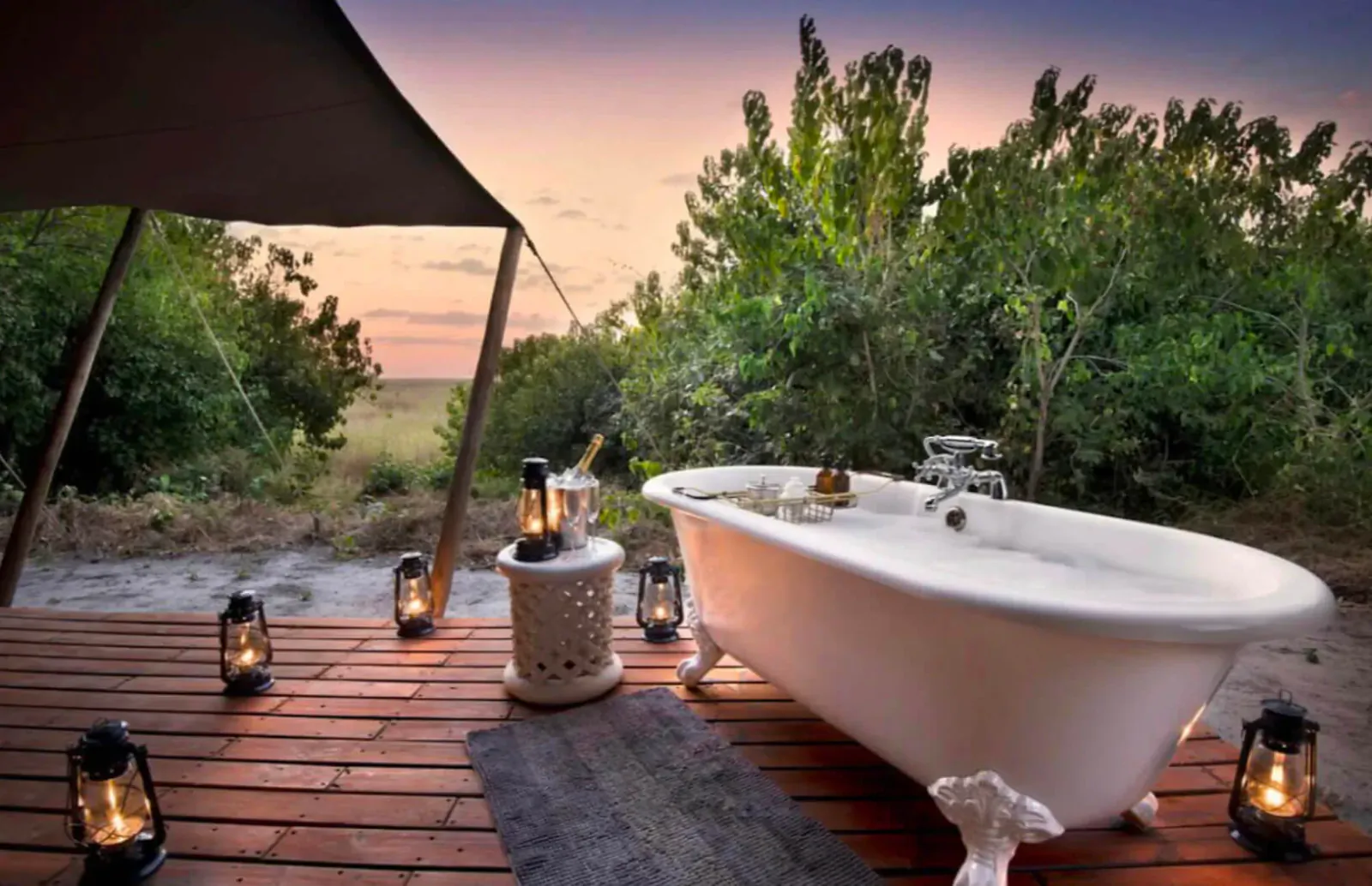 Luxury clawfoot bathtub on wooden deck in African bush camp at sunset, surrounded by lanterns and trees