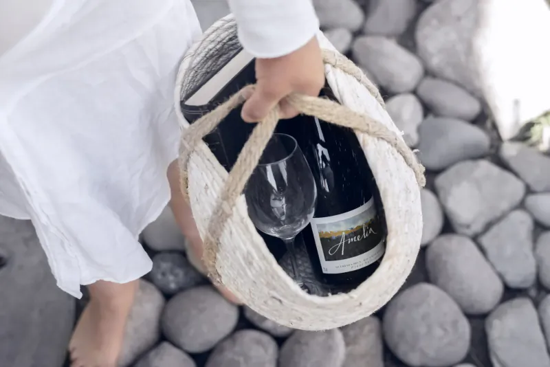 Woman in white dress holding woven basket with Amelia wine bottle and wine glasses on pebble path
