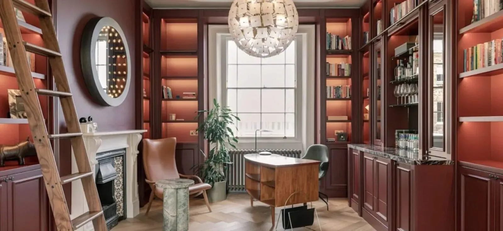Luxurious wood-paneled library with bookshelves, ladder, chandelier, fireplace, desk, chairs, and window.