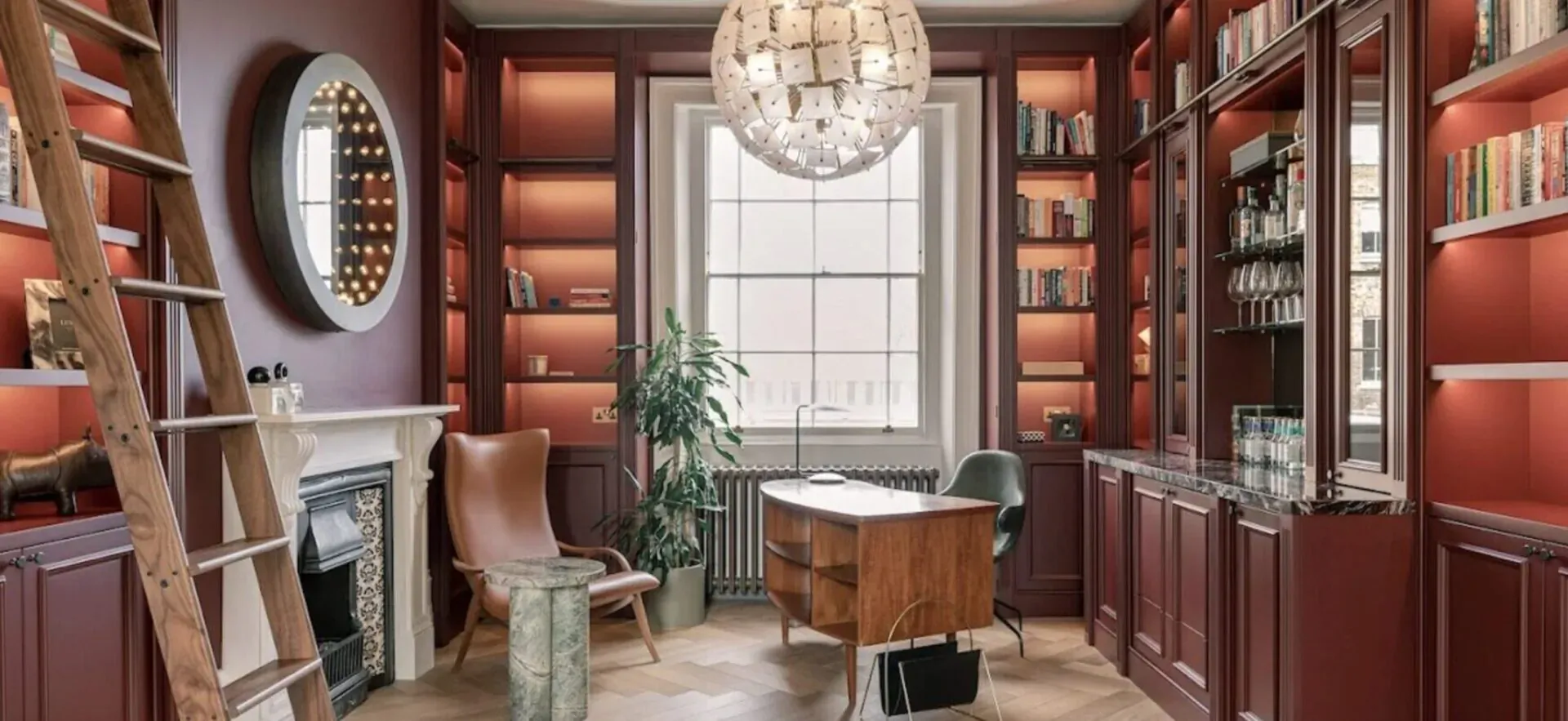 Luxurious wood-paneled library with bookshelves, ladder, chandelier, fireplace, desk, chairs, and window.