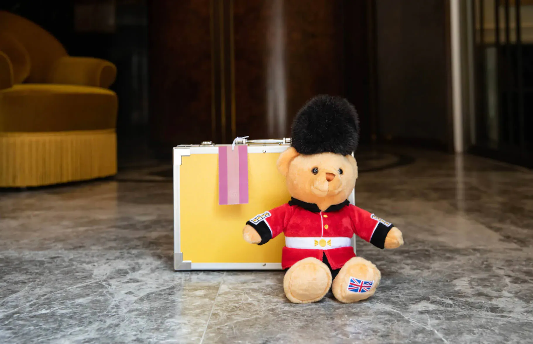 Guardsman teddy bear in red uniform and bearskin hat sits beside yellow suitcase with pink tag in luxury hotel lobby.