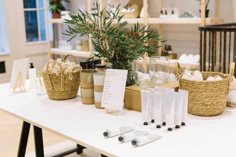 White table with Luxuria skincare products, wicker baskets, glassware, candles, and potted eucalyptus in boutique setting