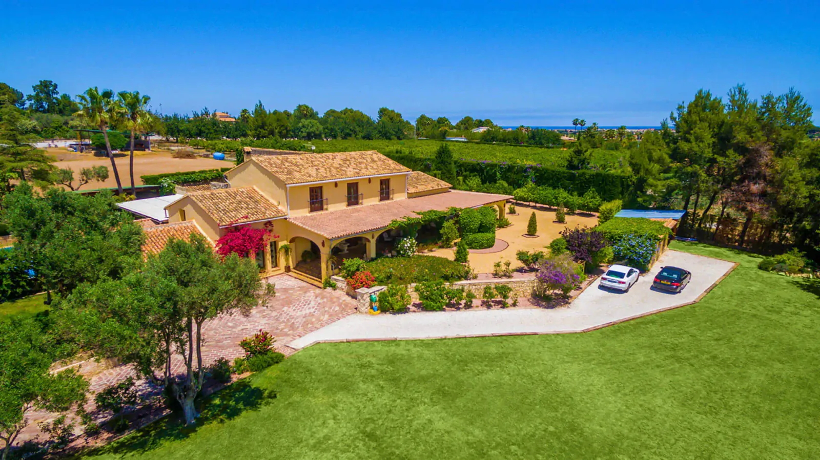Aerial view of a luxurious yellow terracotta-roofed villa with landscaped gardens, palm trees, parked cars, and blue sky.