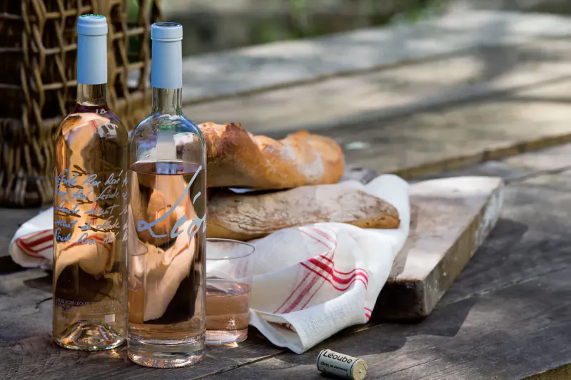 Two Château Léoube rosé wine bottles, baguette bread, and glasses on a wooden table in a picnic basket setting