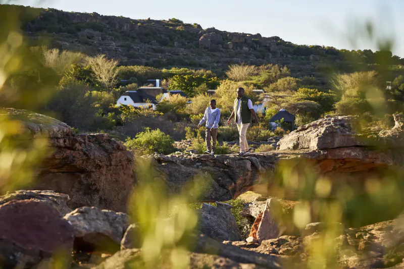 Black couple in casual attire walks hand-in-hand on rocky trail at Bushmans Kloof wilderness reserve, luxury retreat with white cottages in background.