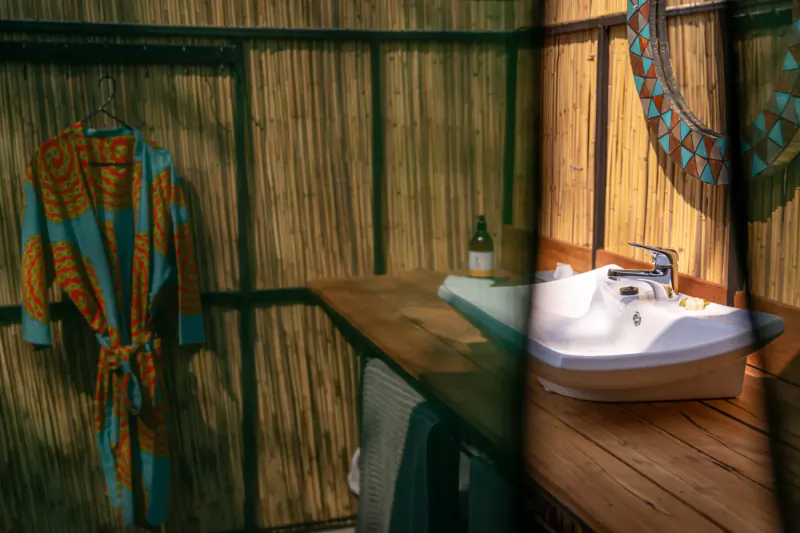 Rustic bamboo-walled bathroom in safari lodge with colorful orange-blue robe hanging by wooden sink and towels