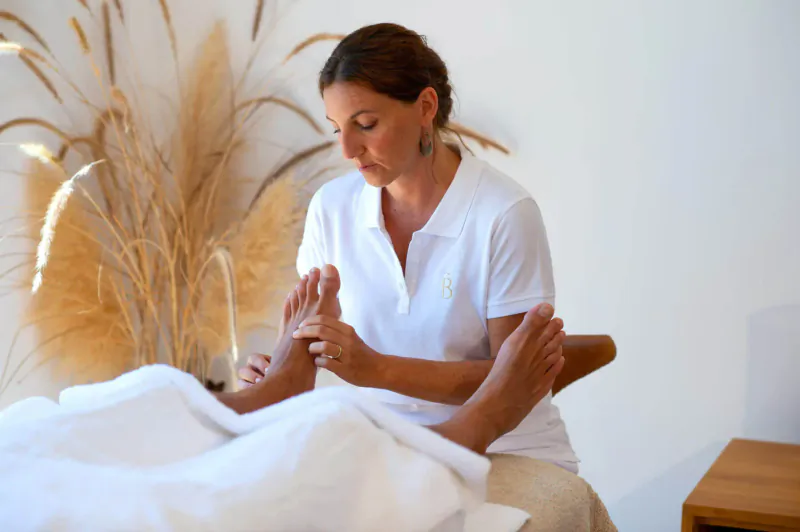 Female therapist in white polo massaging client's feet on white massage bed with pampas grass backdrop