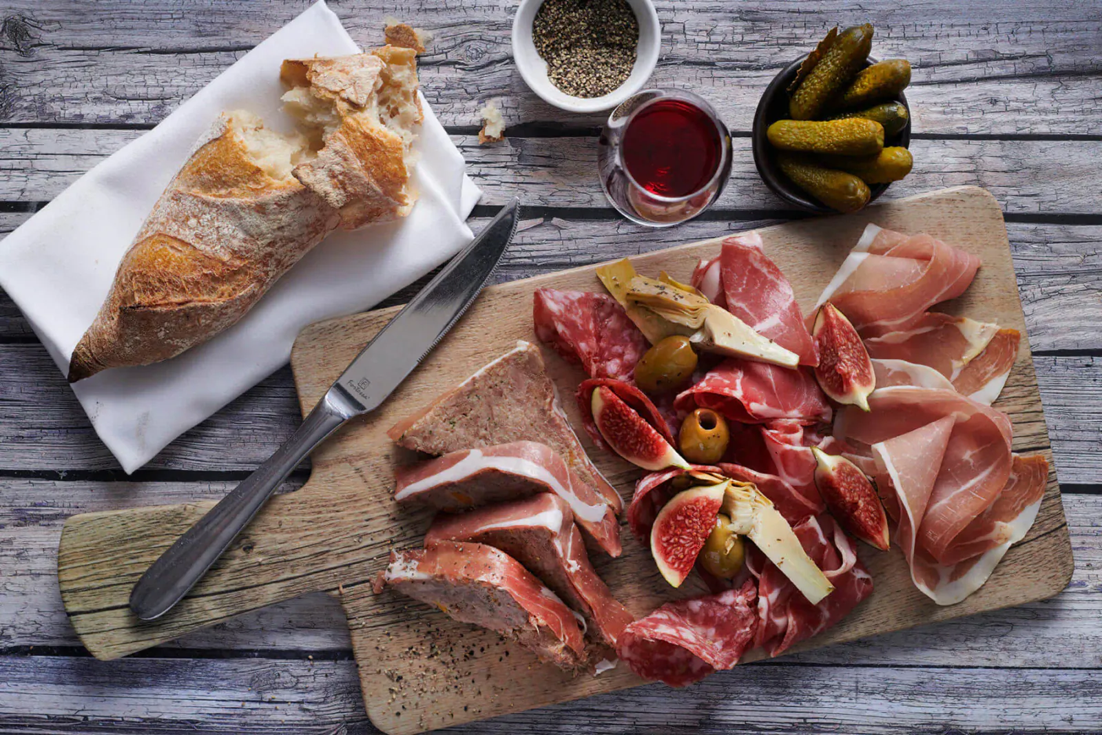 Wooden board with sliced salami, prosciutto, figs, olives, pickles; baguette, knife, pepper, red wine on rustic wood.