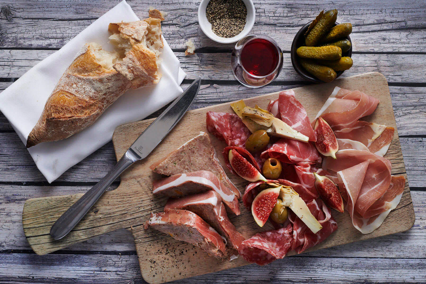 Wooden board with sliced salami, prosciutto, figs, olives, pickles; baguette, knife, pepper, red wine on rustic wood.