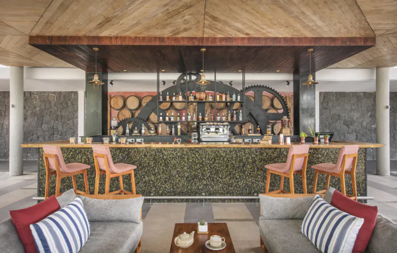 Open-air resort bar with wooden wheel decor, green counter, bar stools, and lounge seating with tea service