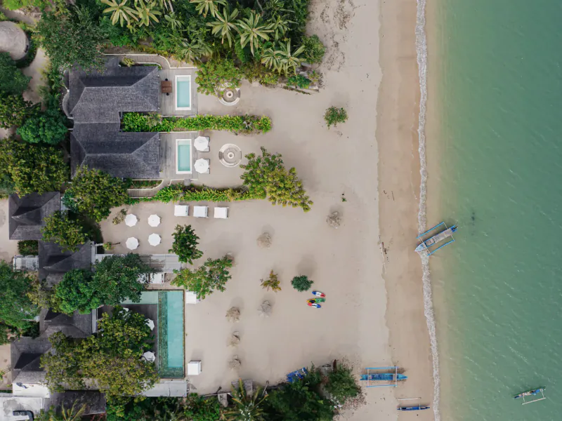 Aerial view of Kuara Lombok resort with thatched villas, infinity pool, sunbeds on sandy beach, and boats in turquoise sea amid palms.