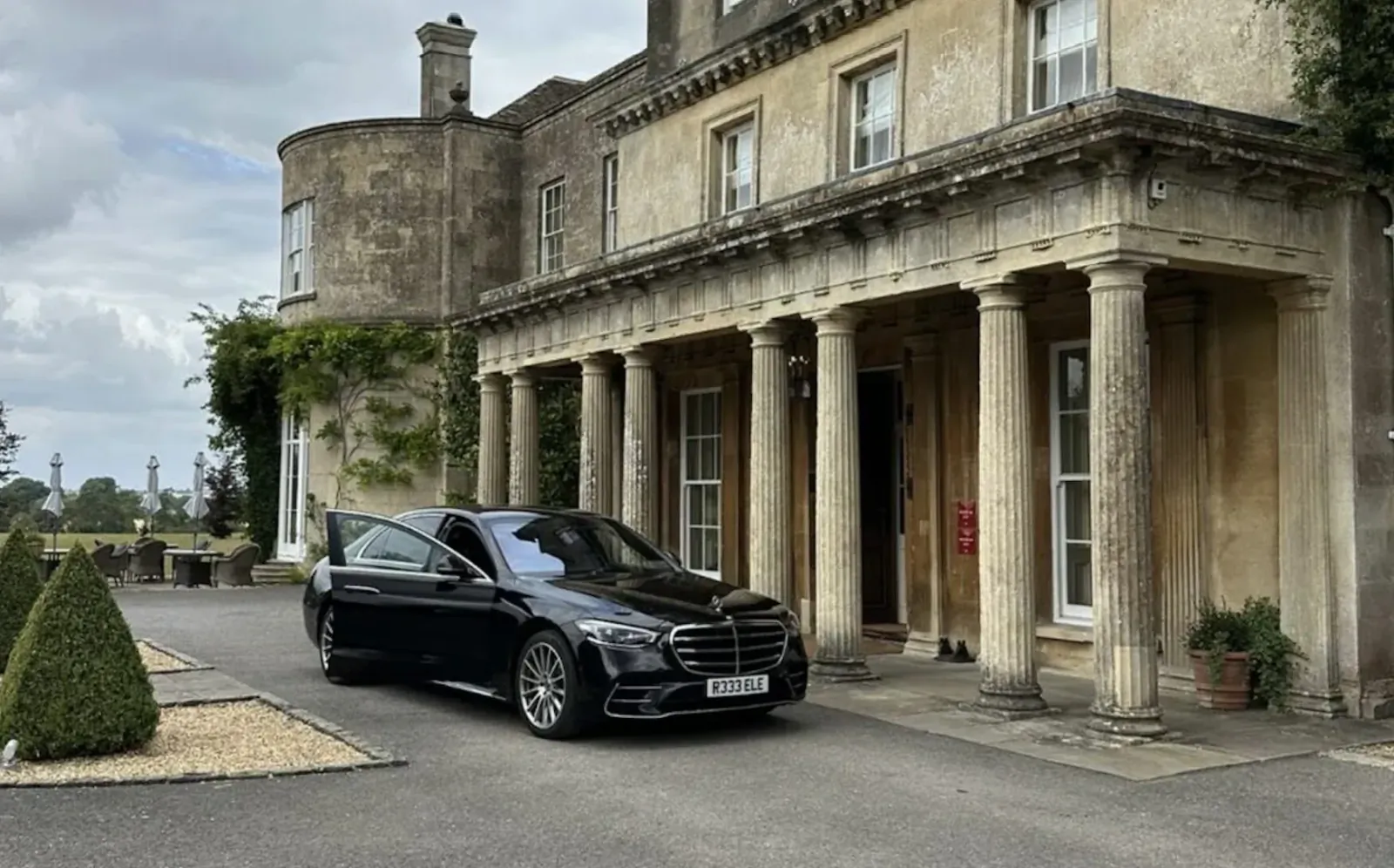 Black Mercedes S-Class luxury car parked in front of elegant stone manor house with columns and manicured gardens.