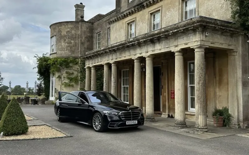 Black Mercedes S-Class luxury car parked in front of elegant stone manor house with columns and manicured gardens.