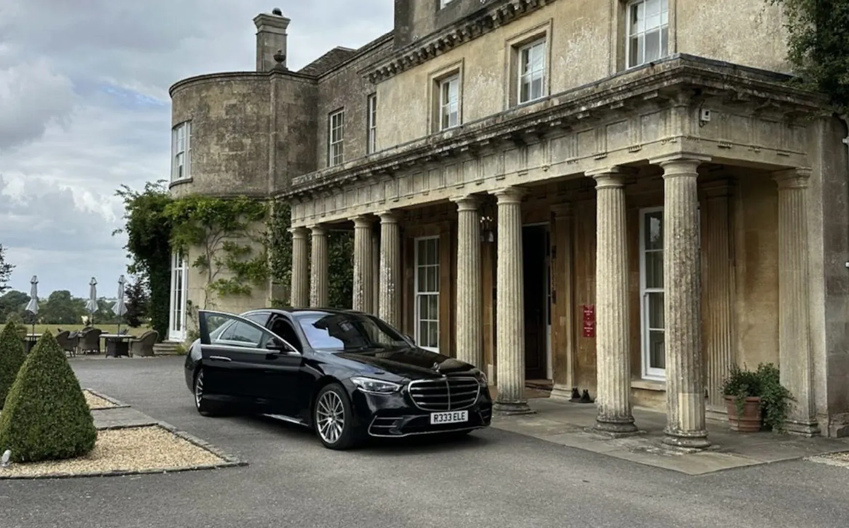 Black Mercedes S-Class luxury car parked in front of elegant stone manor house with columns and manicured gardens.