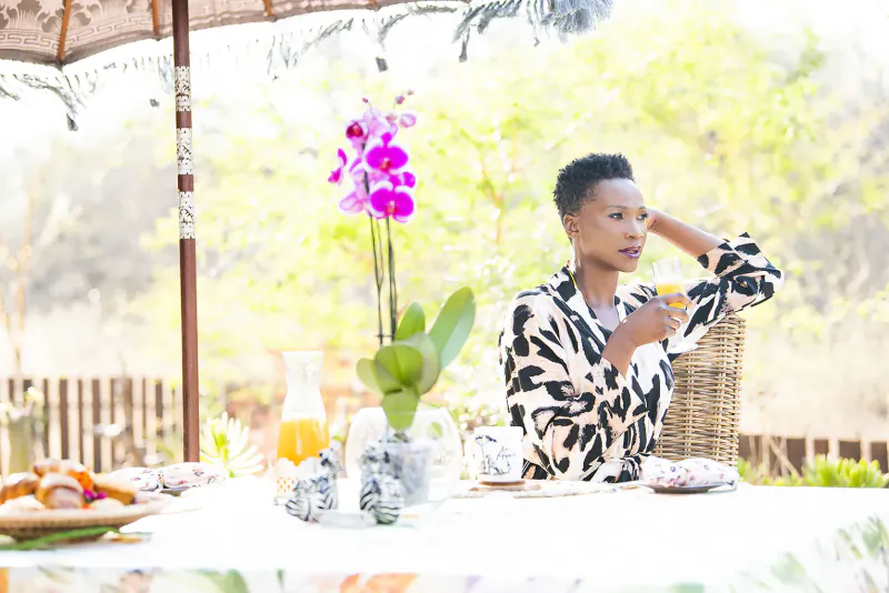 Woman in patterned kimono sips from cup at outdoor table with breakfast, pink orchid, oranges, under safari lodge umbrella