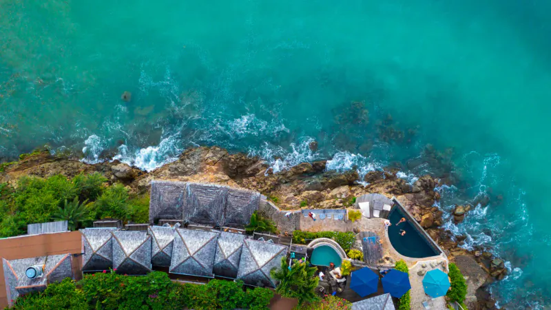 Aerial view of Sheer Rocks resort in Antigua: thatched villas, infinity pools, blue umbrellas on rocky cliffside over turquoise ocean.