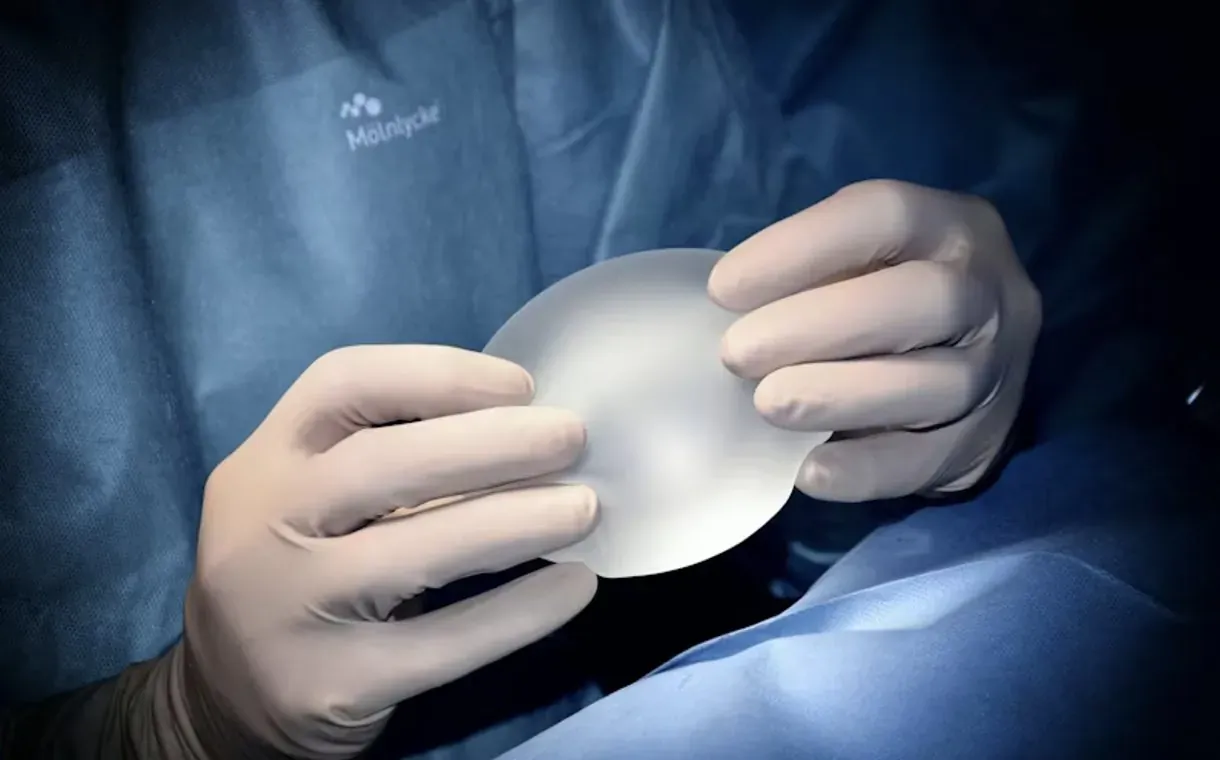 Surgeon's gloved hands holding a clear silicone breast implant in surgery, Motiva logo on scrubs.