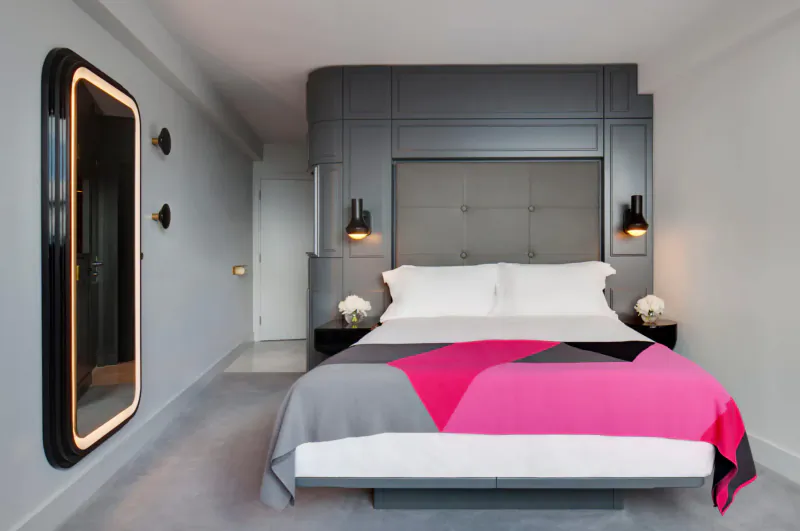 Modern hotel bedroom at Sea Containers London with gray curved walls, lit mirror, pink-gray bedspread, white pillows, and nightstand flowers.