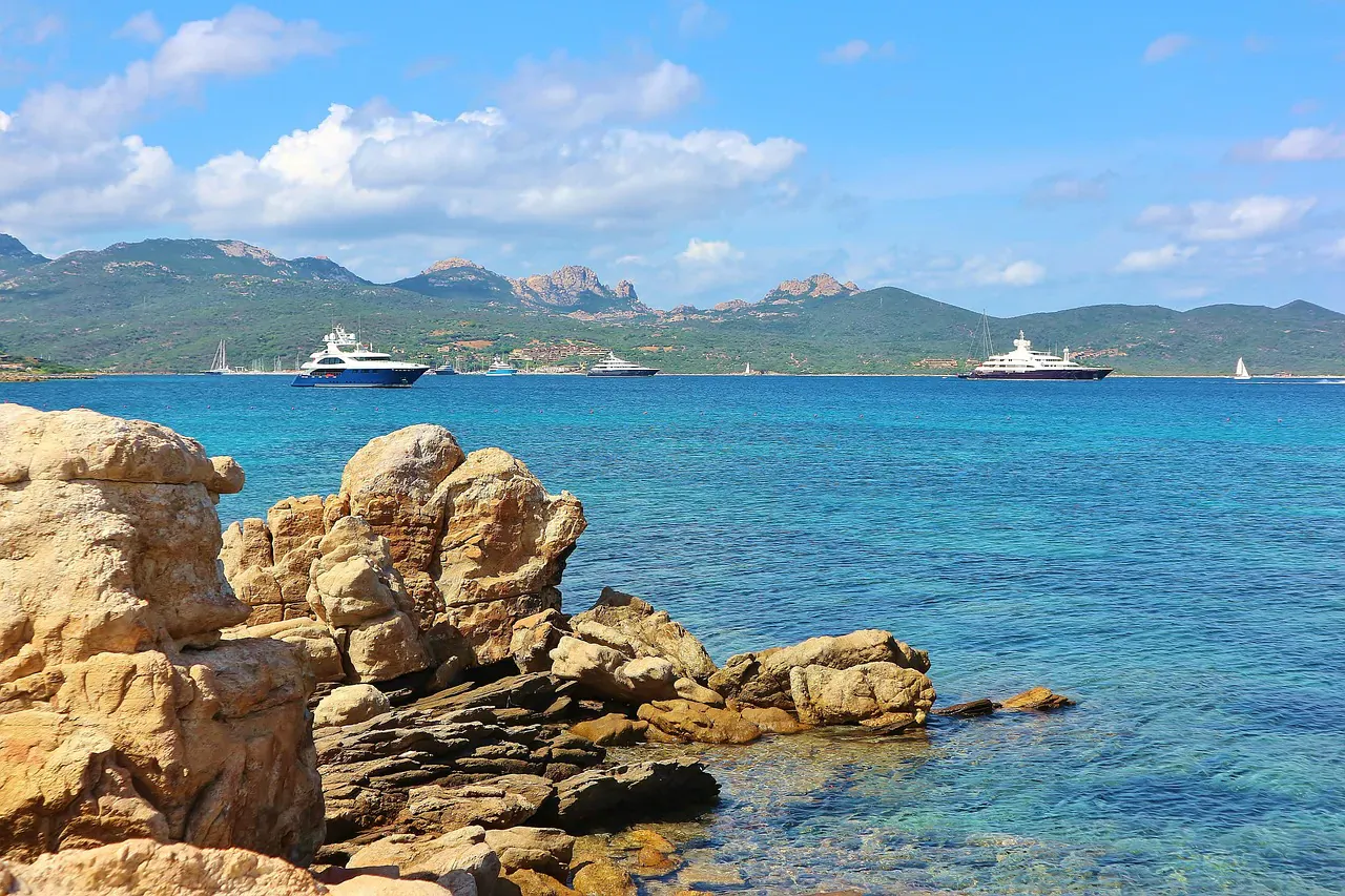 Rocky Mediterranean shoreline with turquoise sea, yachts, cruise ship, and mountains under blue sky, Italy vacation scene