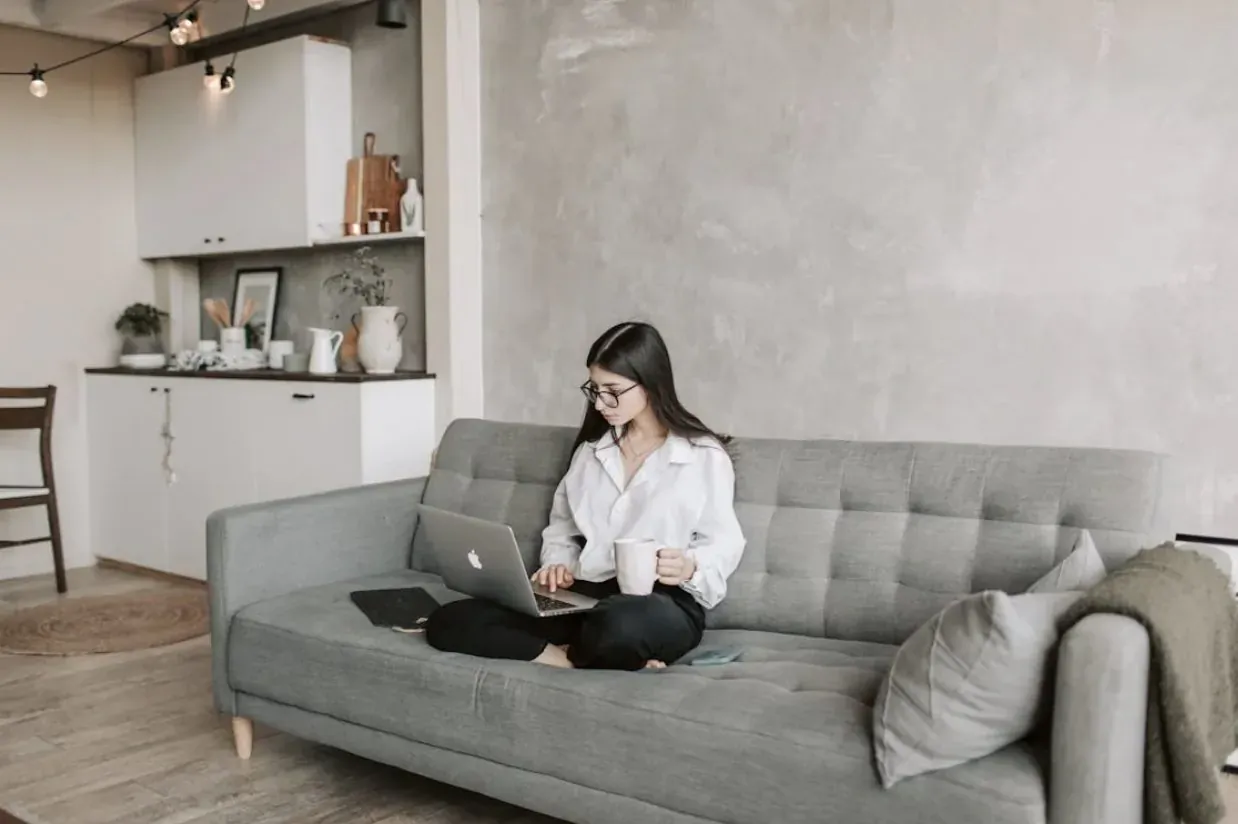 Woman with glasses working on laptop, sitting cross-legged on gray sofa in modern kitchen lounge