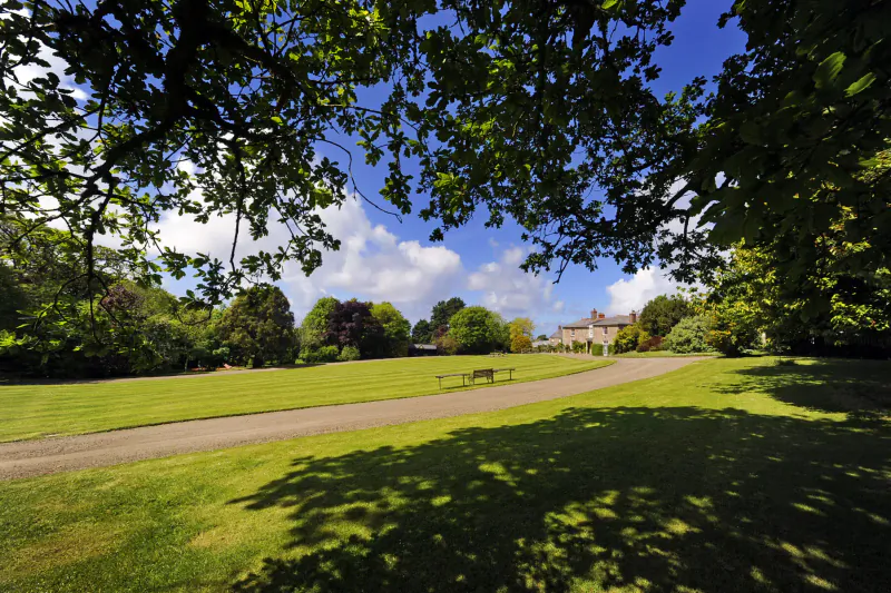 Broomhill Manor, a grand Georgian house in lush Cornish gardens with gravel path and bench under trees