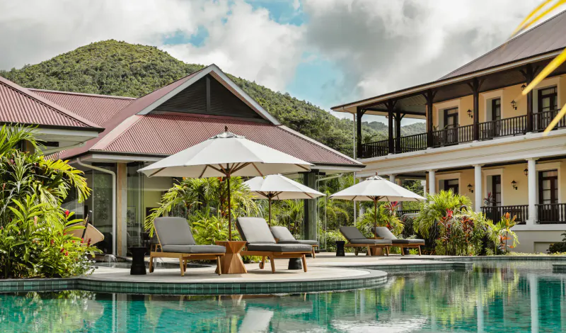 La Cigale Estate in Seychelles: infinity pool with loungers under umbrellas, tropical gardens, red-roofed buildings, mountains beyond.