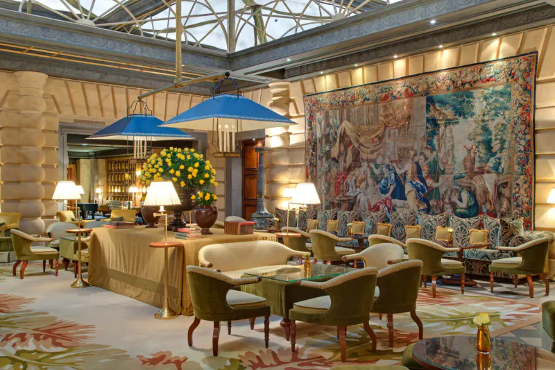 Elegant Hotel Metropole Monte-Carlo lobby with blue umbrellas, yellow flowers, tapestries, lamps, and green armchairs under glass atrium.