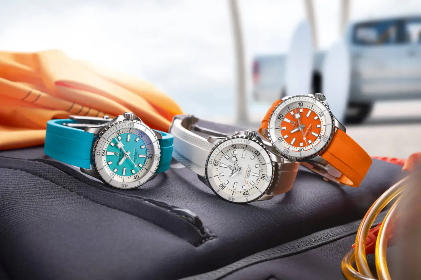 Three Omega Seamaster chronograph watches in turquoise, silver, and orange on black wetsuit near beach truck.