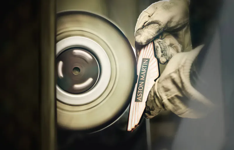 Gloved hands polishing a large wheel, holding Aston Martin wings logo badge