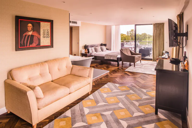 Luxurious hotel suite with beige sofa, king bed, geometric rug, wooden floors, lake view balcony, and portrait painting.