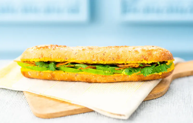 Baguette sandwich with yellow sauce, green herbs, and lettuce on wooden board with napkin