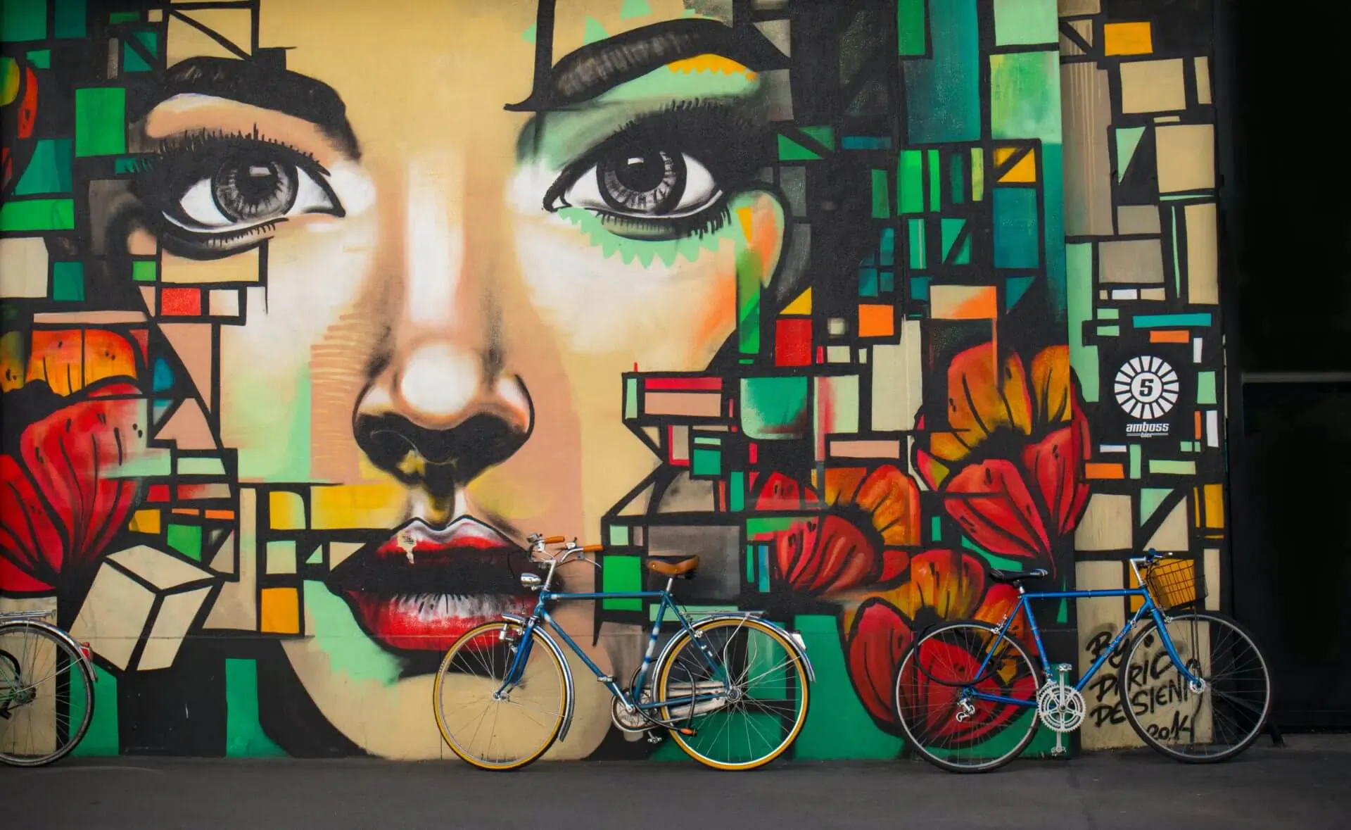 Close-up of a vibrant geometric graffiti mural of a woman's face with green eyes and red lips, colorful bikes parked in front.