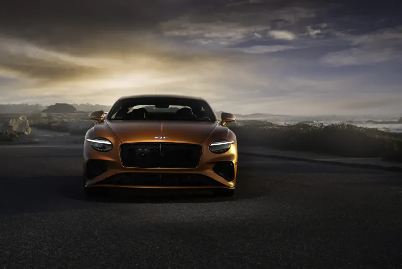 Bentley Continental GT Speed, orange coupe, front view on coastal road at dramatic sunset with ocean backdrop.