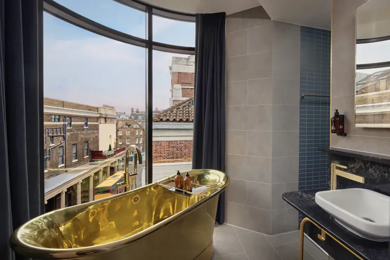 Luxurious hotel bathroom with golden bathtub, city skyline view through curved window, modern sink, Amano Hotel Covent Garden.