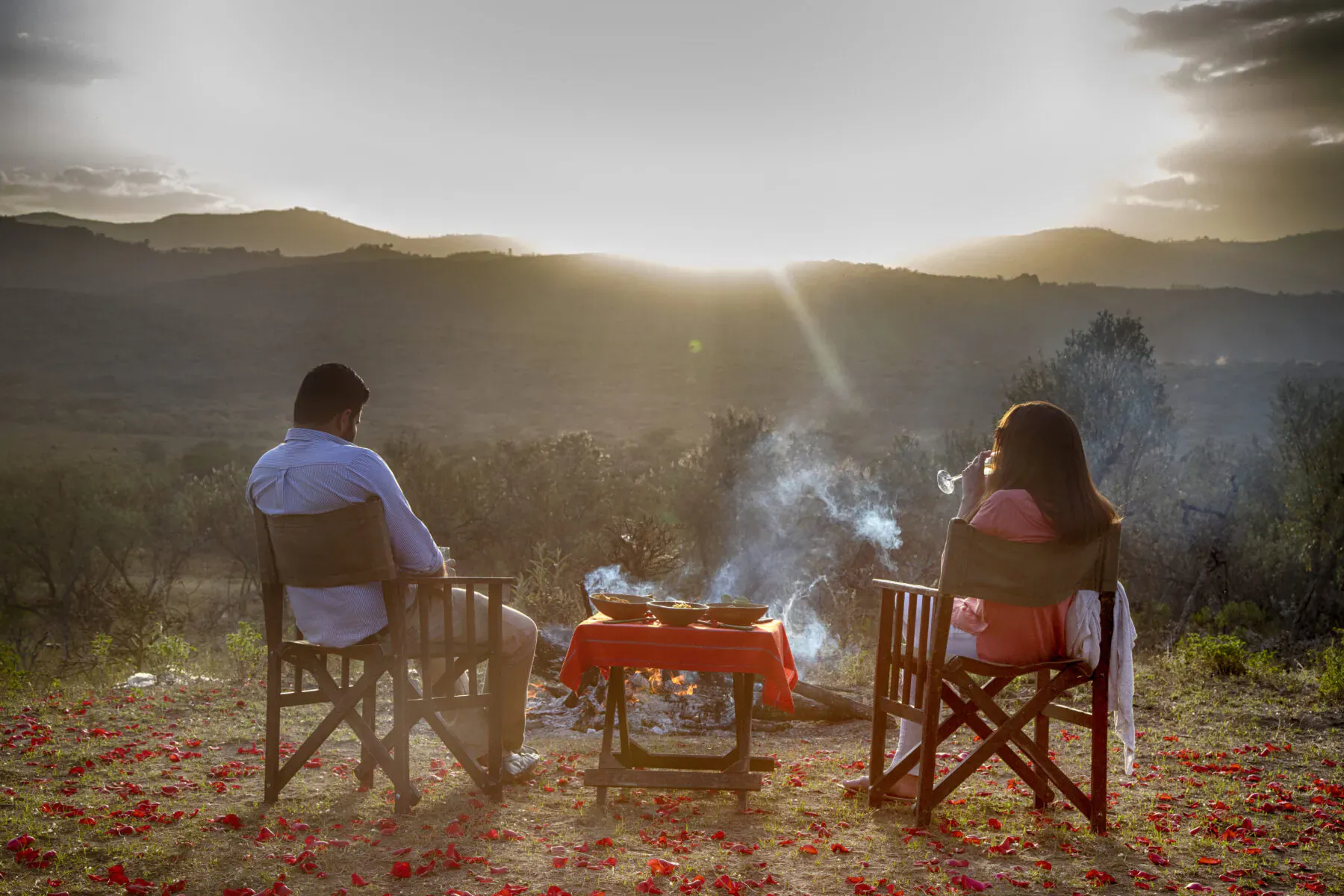 Couple seated at candlelit table by campfire at sunset in Kenyan savanna, Chui Lodge, Lake Naivasha, surrounded by rose petals and mountains.