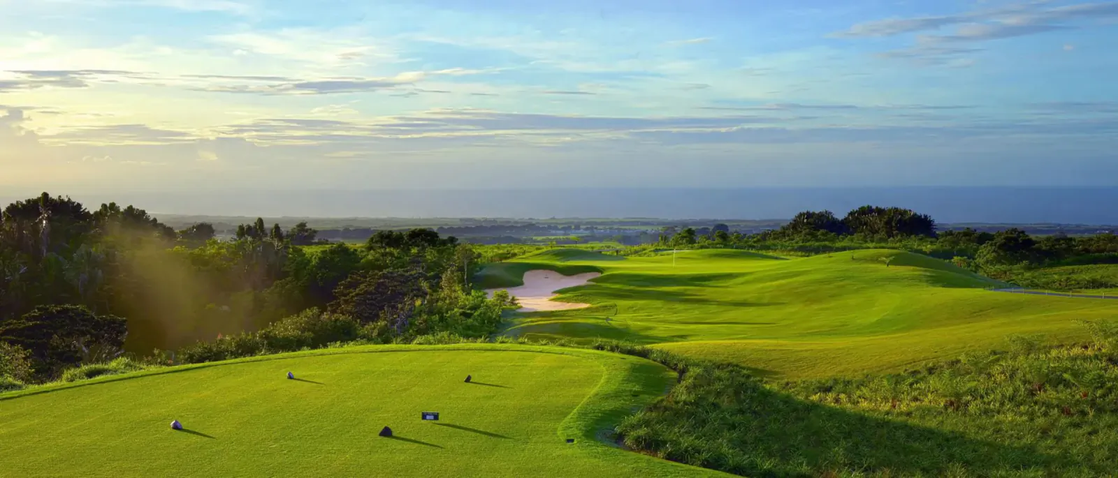 Aerial view of lush green golf course fairways and bunkers at Avalon Golf Estate, Mauritius, with ocean horizon at sunset.