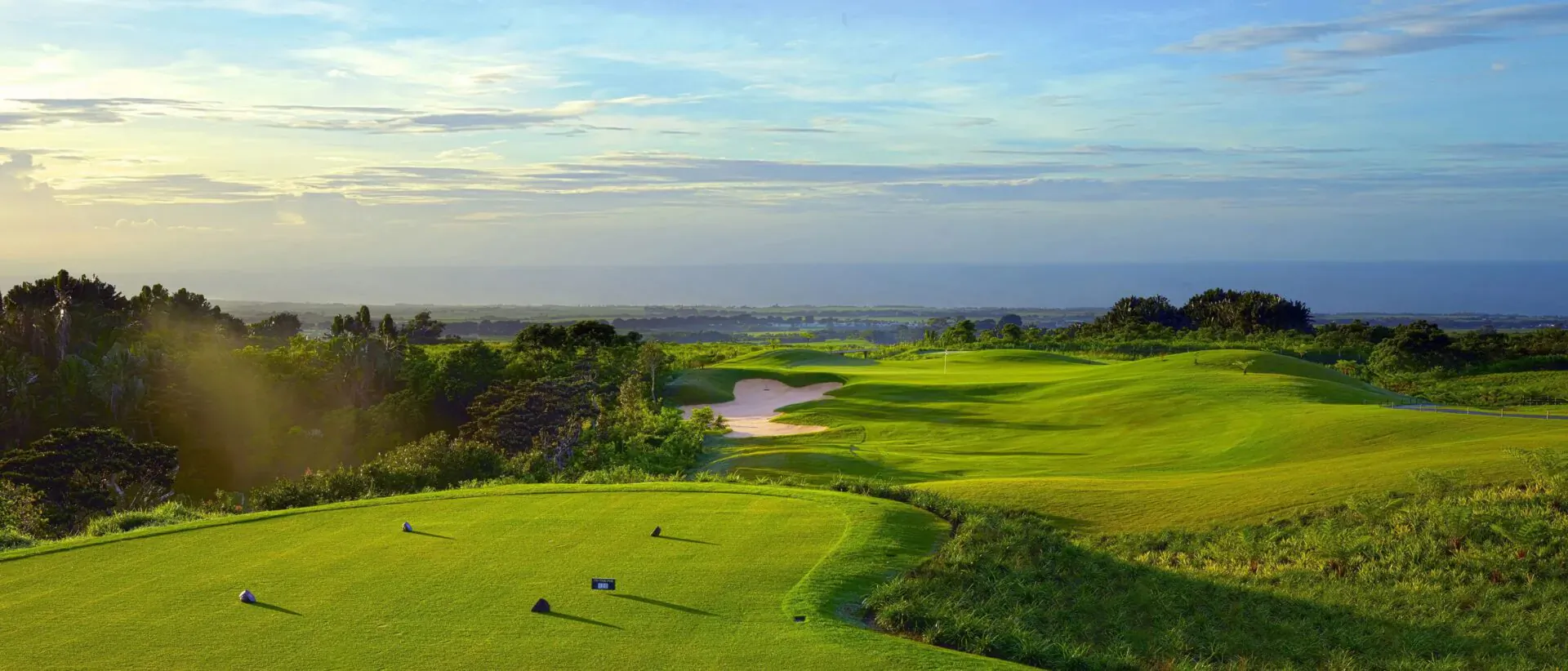 Aerial view of lush green golf course fairways and bunkers at Avalon Golf Estate, Mauritius, with ocean horizon at sunset.