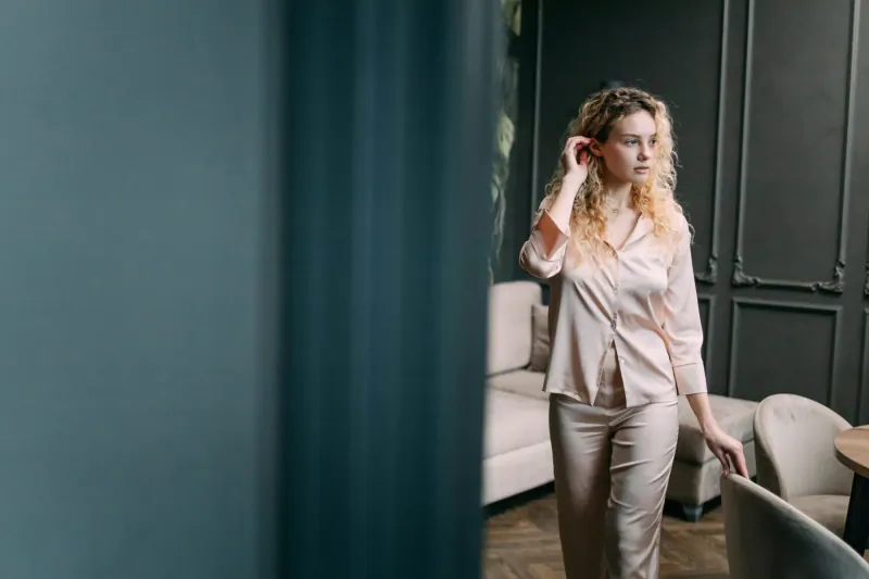Woman with curly hair touching her ear, wearing elegant pink satin pajamas in luxurious room with green curtains and sofa