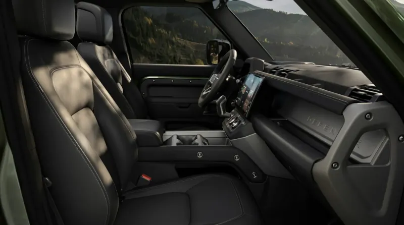 Land Rover Defender interior with leather seats, digital dashboard, steering wheel, viewed from passenger side against mountain backdrop.