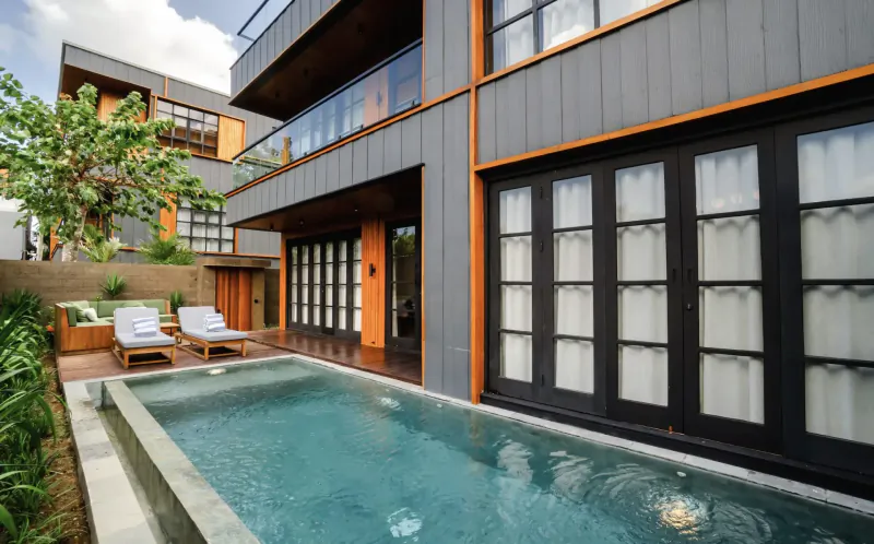 Modern gray villa with infinity pool, lounge chairs, tropical plants, and large glass doors at Secana Beachtown Bali