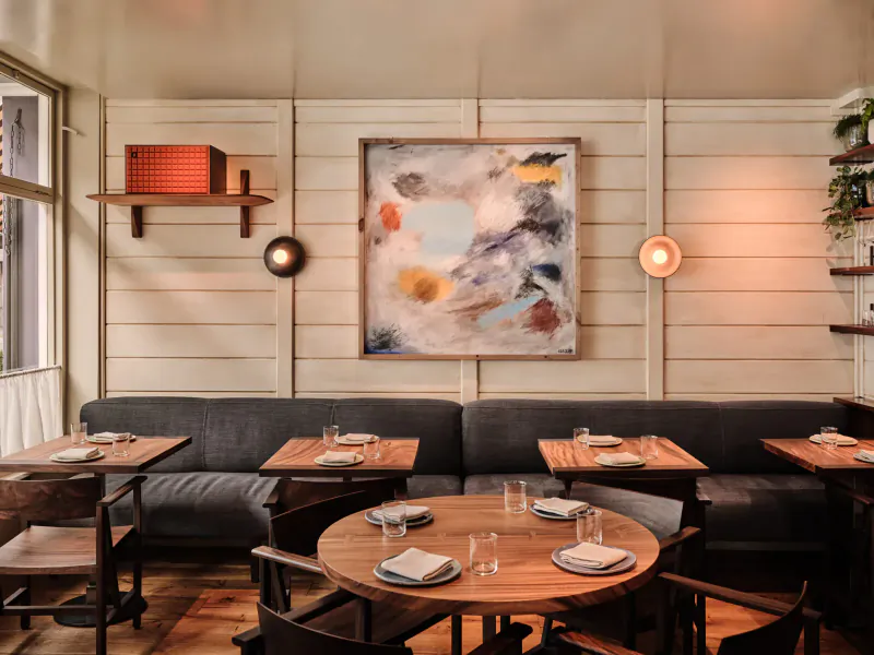 AngloThai restaurant interior: white shiplap walls with abstract painting, plants, round lamps, gray sofas, wooden tables set for dining.