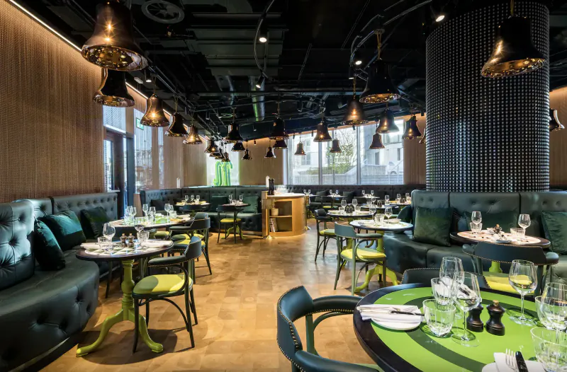 Modern nhow London hotel restaurant with green velvet booths, yellow chairs, round tables, brass pendant lights, and mesh walls.