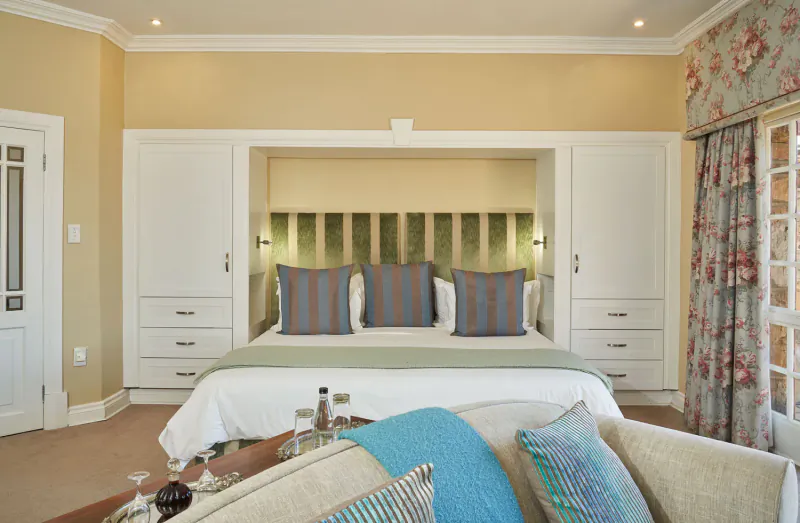 Elegant bedroom with king bed, striped pillows, built-in wardrobe, floral curtains, and wine glasses on nightstand.