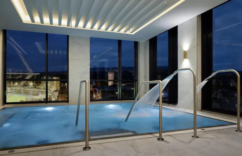 Luxury indoor pool with cascading showers from chrome rails, glowing blue water, large windows showing city night skyline.