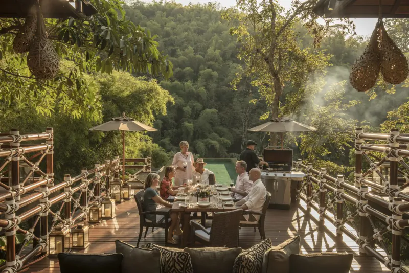 Family dining on a bamboo deck at Four Seasons Thailand resort, grilling amid lush jungle and pool views.