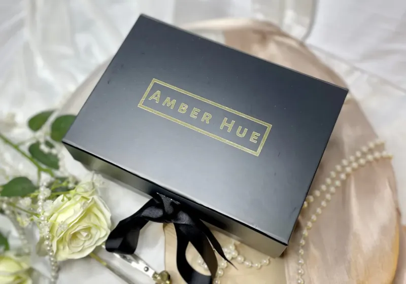 Black 'Amber Hue' box with black ribbon on pink satin, beside white roses, pearl necklace, and wine glass.