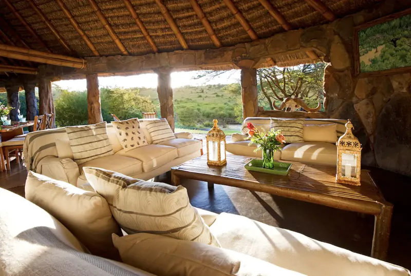 Cozy lounge at El Karama Lodge, Kenya: beige sofas, wooden table with flowers, lanterns, thatched roof, savanna view.